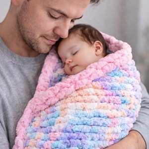 Könnte beinhalten: Eine grob gestrickte Babydecke mit einem bunten Farbmuster aus zartem Rosa, Hellblau, Lavendel und Weiß. Die Decke weist ein dickes, strukturiertes Schlingenmuster auf und ist um einen schlafenden Säugling gewickelt.