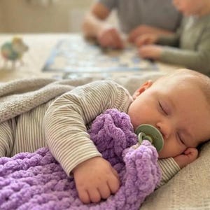 Könnte beinhalten: Ein schlafender Säugling trägt ein langärmeliges Oberteil mit dünnen beigen und weißen Streifen und hält eine weiche, grob gestrickte violette Decke. Der Säugling hat einen hellgrünen Schnuller im Mund und liegt auf einem strukturierten Stoff.
