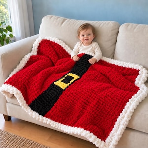 May include: A chunky knit blanket featuring a red base with a wide black horizontal stripe and a yellow rectangular buckle design, finished with a thick white pom-pom border. The blanket is draped over a light-coloured sofa.