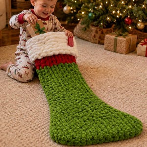Puede incluir: Un calcetín navideño de punto grueso con cuerpo verde, una banda roja y un puño blanco grueso. El calcetín está hecho de hilo suave y texturizado y se coloca sobre una alfombra de color claro, mostrando su diseño acogedor y grande.