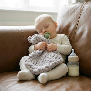May include: A sleeping infant wearing a long-sleeved cream-colored shirt and pants, holding a chunky knit light gray blanket. The infant has a sage green pacifier in their mouth. A clear plastic baby bottle filled with milk sits nearby.