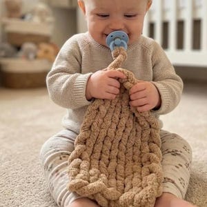 May include: A beige chunky knit security blanket with a soft, textured surface held by a baby wearing a light beige long-sleeve jumper and patterned leggings, with a light blue dummy in their mouth, showcasing a cosy, tactile design.