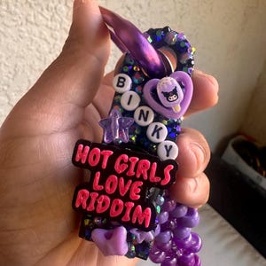 May include: A purple and black glittery keychain featuring white letter beads spelling BINKY, a purple heart charm with a character graphic, a purple star, and a black rectangular plaque with pink text reading HOT GIRLS LOVE RIDDIM.