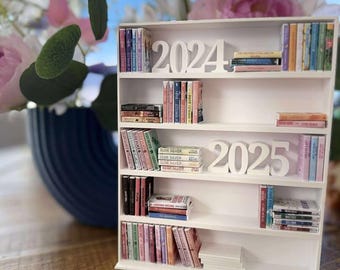 Peut inclure: Une étagère blanche à quatre niveaux, remplie de livres de poche colorés. L'étagère supérieure porte les chiffres 2024, la troisième étagère porte les chiffres 2025.