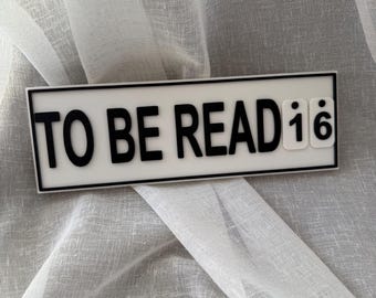 May include: A rectangular sign with the words "TO BE READ" in bold black letters on a white background. The number "16" is at the end of the text. The sign is framed in black and rests on a light gray, textured fabric.