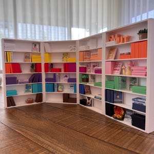 Puede incluir: Una estantería blanca en miniatura llena de libros coloridos, objetos decorativos y una pequeña planta en maceta. La unidad está dispuesta en zigzag, con el número "25" visible en una sección.