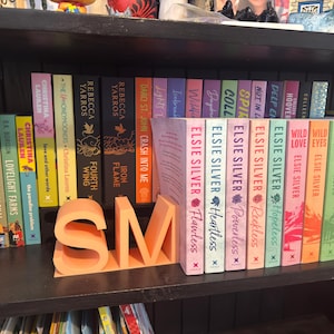 May include: A bookshelf filled with colorful books, including titles by Elsie Silver, Rebecca Yarros, and Christina Lauren. A peach-colored "SM" bookend sits in front of the books, adding a decorative touch to the shelf.