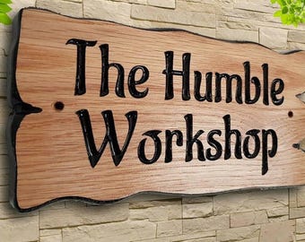 Puede incluir: Un letrero de madera con el texto "The Humble Workshop" pintado en letras negras. El letrero tiene un aspecto rústico y desgastado y está montado en una pared de ladrillo.