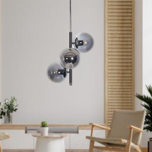 Pode incluir: Uma luminária pendente moderna com três esferas de vidro prateadas penduradas em uma estrutura de metal preto. As esferas são dispostas em forma de escada, com a esfera maior na parte superior e a menor na parte inferior.