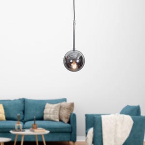 Könnte beinhalten: Eine Chrom-Pendelleuchte mit einem runden, verspiegelten Glasschirm. Die Leuchte hängt an einer weißen Decke und beleuchtet ein Wohnzimmer mit einem blauen Sofa und einer weißen Wolldecke auf einem Stuhl.