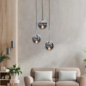 Peut inclure: Trois lampes suspendues chromées avec des abat-jours en verre fumé suspendues au plafond. Les lampes sont suspendues au-dessus d'un canapé brun clair avec deux coussins.