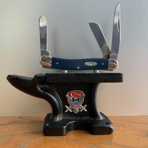 May include: A navy blue Case pocket knife with three open blades rests on a black anvil. The knife has a silver blade and a navy blue handle. The anvil has a Case logo with an eagle and crossed axes.