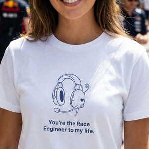Puede incluir: Una camiseta blanca con un diseño de auriculares azules y el texto "You're the Race Engineer to my life". La imagen muestra a una persona en un entorno de carreras, con otras personas y edificios al fondo.