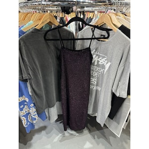 May include: A dark purple, sparkly spaghetti strap dress hangs on a black hanger. The dress is surrounded by various t-shirts on wooden hangers, all displayed on a clothing rail. The t-shirts are in shades of grey and blue.