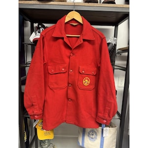 May include: A vibrant red wool shirt jacket with two chest pockets and a button-down front. The jacket has a Boy Scouts of America emblem on the right chest. The jacket is hanging on a wooden hanger.