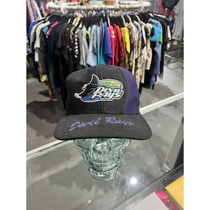 Peut inclure: Casquette de baseball noire avec un panneau latéral violet et une visière plate. La casquette affiche le texte "Tampa Bay Devil Rays" dans un logo stylisé à l'avant et les mots "Devil Rays" brodés sur la visière. La casquette est présentée sur un crâne en verre.