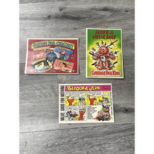 May include: Three vintage Garbage Pail Kids trading cards. One card reads "GARBAGE PAIL CIRCUS!" with cartoon characters. Another card says "HAVE A NICE DAY" with a character holding weapons. The third card says "BAZOOKA JERK" with comic strip art.