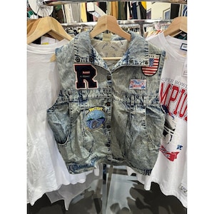 Może przedstawiać: Dżinsowa kamizelka w stylu acid wash z naszywkami, w tym dużą literą "R" i amerykańską flagą. Kamizelka ma zapięcie na guziki i dwie kieszenie. Wisi na drewnianym wieszaku.