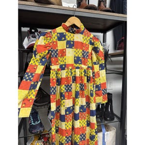Peut inclure: Une robe vintage patchwork avec un col montant et des manches longues. La robe présente un motif coloré de carrés jaunes, rouges et bleus, avec de petits détails floraux et des visages souriants. La robe est suspendue à un cintre en bois.