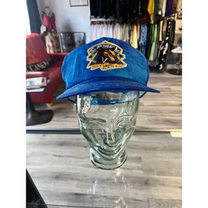 Può includere: Cappellino da camionista blu con visiera curva e toppa con la scritta "CAMEL" e "Smooth Character". Il cappello è esposto su una testa di vetro.