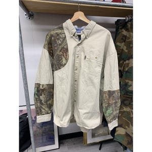 May include: A long-sleeved button-down shirt with a beige body and camouflage patterned sleeves and shoulder accents. The shirt has a button-down collar and a single chest pocket. It is hanging on a wooden hanger.