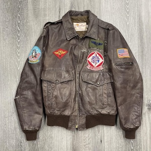 May include: A brown leather bomber jacket with multiple patches, including a Tomcat patch, an American flag, and other military insignia. The jacket has two front pockets, a ribbed hem, and a collar. The jacket is a vintage style.