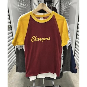 May include: A maroon and gold raglan t-shirt with the word "Chargers" in yellow script. The shirt has a yellow collar and sleeves. The shirt is hanging on a wooden hanger.