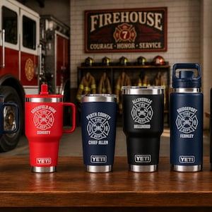 May include: Six stainless steel YETI tumblers and bottles in navy, red, and black, each featuring a white fire department emblem with specific location and name text. The drinkware is arranged in a row on a wooden surface in a fire station.