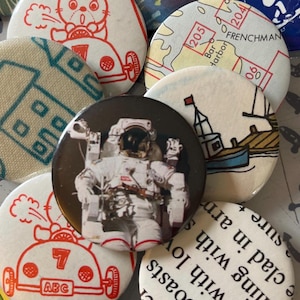 May include: A collection of round pinback buttons with various designs. One button features an astronaut in a spacesuit. Other buttons display a map, a boat, a car, and cartoon characters. Some buttons have text.