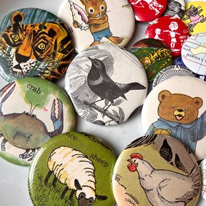 Op de afbeelding: Een verzameling kleurrijke, ronde buttons met illustraties van dieren. Ontwerpen omvatten een tijger, konijn, krab, schaap, beer en een kraai. Sommige buttons hebben tekst. De buttons zijn in verschillende stijlen.