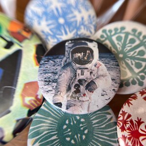 May include: A collection of round buttons with various designs. One button features an astronaut on the moon. Other buttons have blue, green, and red patterns. The buttons are likely for decorative purposes or as accessories.