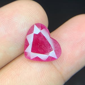 May include: A heart-shaped, faceted gemstone in a vibrant shade of red. The gemstone has a polished surface with multiple facets that reflect light, creating a sparkling effect. The gem is held between fingers.