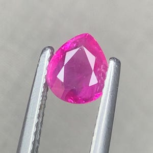 Puede incluir: Una gema facetada en forma de pera de un tono rosa intenso, sostenida firmemente por unas pinzas de metal plateado. La piedra presenta una superficie clara con múltiples facetas que refleja la luz, mostrando su color brillante.