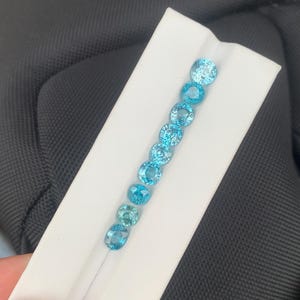 May include: A collection of round and oval-cut, light blue gemstones displayed on a white card. The gemstones vary slightly in shade, ranging from a pale turquoise to a deeper aqua. The arrangement highlights the gems' clarity and color.