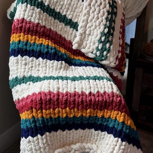 May include: A chunky knit throw blanket with a striped pattern in shades of red, yellow, blue, and green. The blanket is made of a soft, fluffy yarn and is draped over a chair.