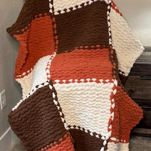 May include: A chunky knit throw blanket featuring a patchwork design of large squares in solid cream, dark brown, and burnt orange colors. The blanket is constructed from thick, soft yarn loops with visible stitching connecting each square section.