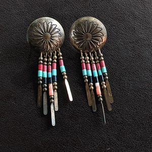 Könnte beinhalten: Silberfarbene Ohrringe mit einem kreisförmigen Design mit Sonnenstrahlen. Sie haben einen Perlenbesatz in Rot, Türkis, Schwarz und Koralle, mit kleinen Metallakzenten und länglichen Metalltropfen. Ein auffälliges Accessoire.