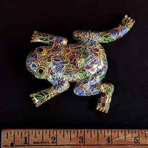 May include: A gold-toned frog brooch with intricate floral enamel detailing in shades of blue, purple, red, and green. The frog's body is adorned with a detailed pattern, and it has black eyes. The brooch is set against a dark background.