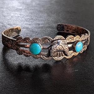 Puede incluir: Una pulsera de puño plateada con piedras turquesas y un diseño central detallado. La pulsera presenta un patrón decorativo en los bordes. La pulsera es una pieza de joyería.
