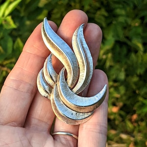 May include: A silver-toned brooch with a textured, abstract design. The brooch features curved, leaf-like shapes that intertwine, creating a dynamic and elegant accessory. The metal has a brushed finish, adding depth and visual interest.