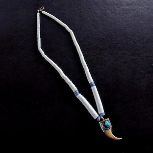 May include: A white beaded necklace with blue accents and a pendant. The pendant features a silver-colored setting with a turquoise stone and a claw-shaped element. The necklace is displayed on a black surface.