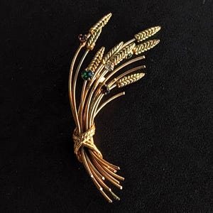 Puede incluir: Broche dorada con forma de espiga de trigo, adornada con pequeñas piedras preciosas de colores. La broche presenta un nudo tejido detallado en la base y delicados tallos curvos. Las piedras preciosas son en tonos verdes, morados y claros.