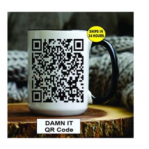May include: White ceramic mug with a black handle and rim, featuring a large black and white QR code design. The text "DAMN IT QR Code" is printed below the QR code. The mug is set on a wooden surface.