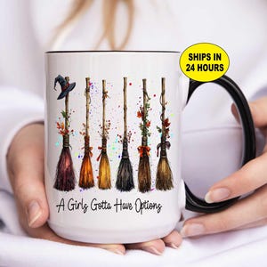 May include: White ceramic mug with a black handle and rim, featuring a design of six colorful witch brooms with autumn leaves and a witch hat. The text "A Girls Gotta Have Options" is printed on the mug.