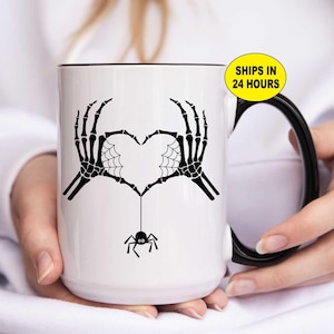 May include: White ceramic mug with a black rim and handle. The mug features a black skeleton hand design forming a heart shape with a spider and web detail. The text "SHIPS IN 24 HOURS" is on a yellow oval.