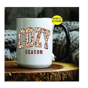 May include: White ceramic mug with a black handle and rim. The word "COZY" is printed in a floral pattern, and the word "SEASON" is printed below in a brown font. The mug is sitting on a wooden surface.