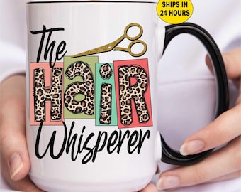 May include: White ceramic mug with a black handle and rim. The mug features the text "The Hair Whisperer" in a script font, with the word "Hair" in leopard print letters. A gold-colored pair of scissors is above the word "Hair".