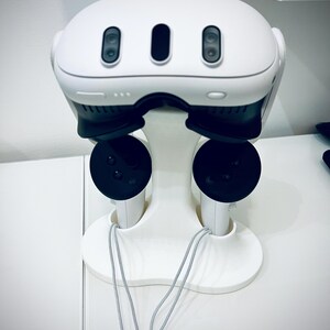 May include: A white VR headset with three black lenses and a matching white stand. The stand holds two black controllers with gray lanyards. The headset and stand are on a white surface.