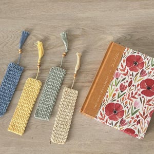 May include: Five crocheted bookmarks with wooden beads and tassels. The bookmarks are in blue, yellow, green, and beige. The bookmarks are displayed on a wooden surface next to a closed book with a floral design and the text "The New Testament -  Faithlife".