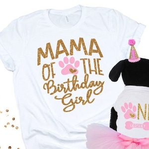 May include: A white t-shirt with gold glitter text reading MAMA OF THE BIRTHDAY GIRL and a pink paw print. Beside it, a black plush toy wears a white shirt with gold glitter text reading ONE, a pink paw print, and a pink tutu with a party hat.